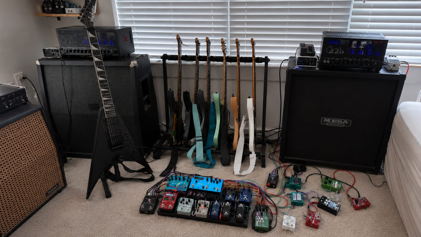 The full stereo guitar rig: a rack of headless Strandberg guitars flanked on the left by a black Jackson King V leaning on a 4x12 cab with a blue-lit head on top, and on the right a PRS MT15 head glowing blue atop a Mesa/Boogie 4x12, with a large pedalboard and floor pedals wired in colorful patch cables in the foreground.